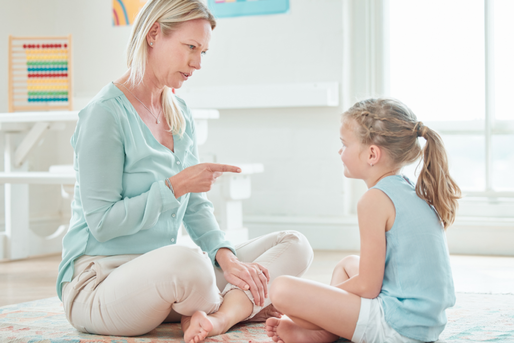 Une maman assise sur le soleil en tailleur avec sa fille assise de la même manière en face d'elle. La maman pointe le doigt vers sa fille pour instaurer une limite avec une explication bienveillante.