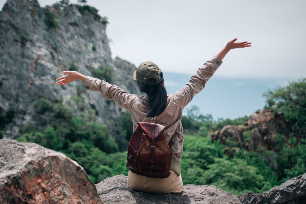 Une femme qui prend du temps pour elle en pleine nature assise sur un rocher face à la verdure et à la mer avec les bras écartés et en l'air pour exprimer la joie de ce moment.