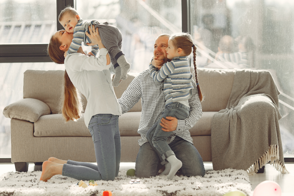 Un moment de complicité en famille. La maman porte le bébé en l'ai en lui déposant un bisou sur la joue et le papa porte sa fille déjà plus grande et lui fait un câlin. Cette photo résume bien la complicité des moments simple en famille. 